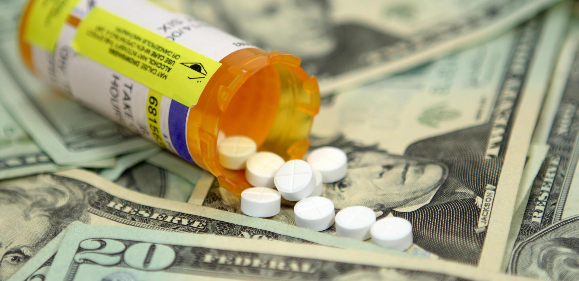 Prescription pill bottle spilling white tablets onto U.S. twenty-dollar bills, symbolizing the cost of medications and drug pricing issues.